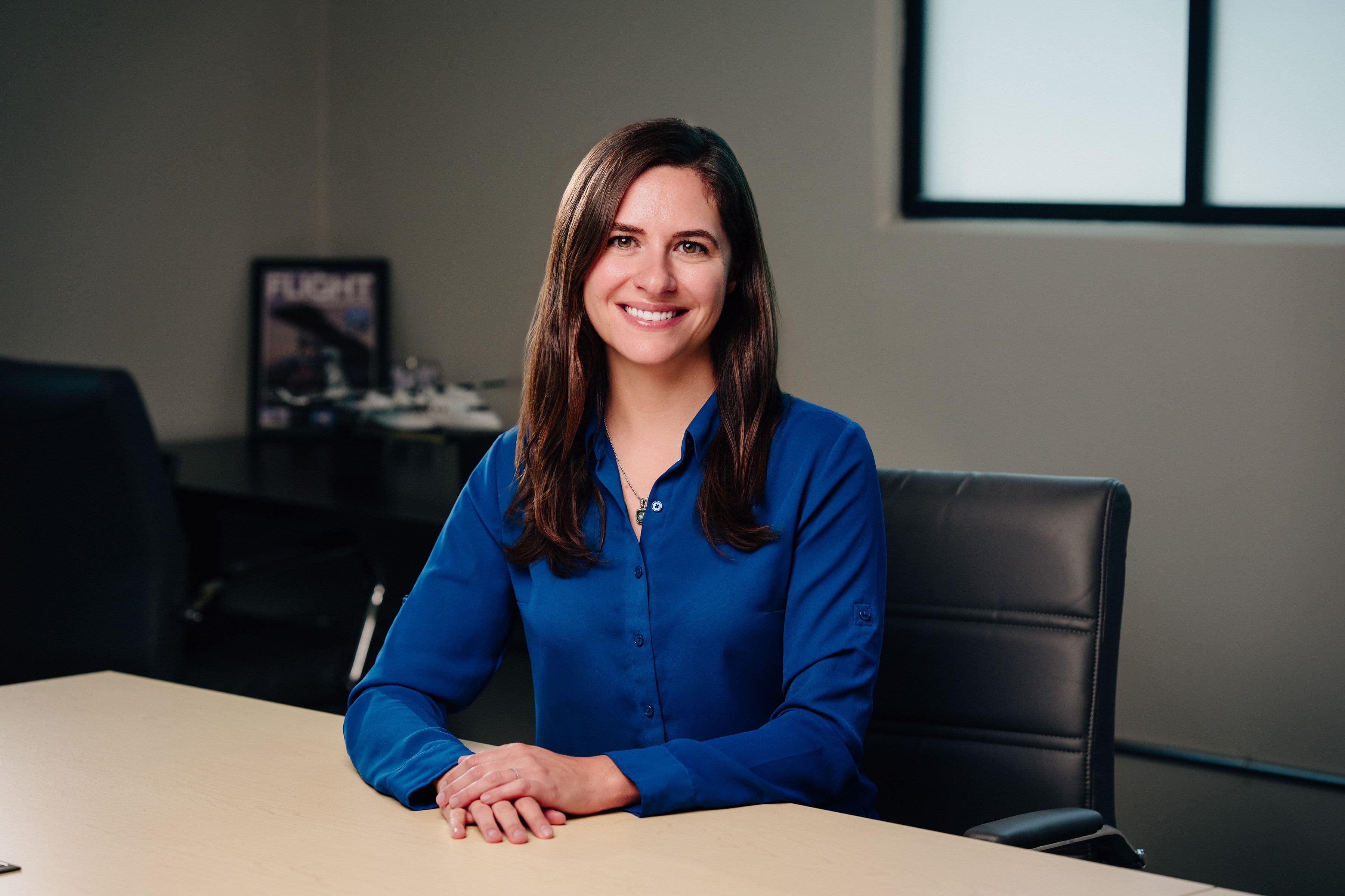 A smiling woman with long dark hair, wearing a blue button-up shirt, sitting at a conference table with her hands clasped in front of her in an office setting.